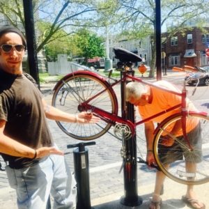 A person pointing to another person who is working on a red bike at a local fix-it station