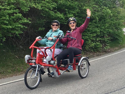 Two women riding an adaptive cycle and waving 