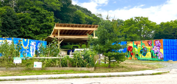 The Big Blue Bike Barn campus. Community garden in foreground, shipping containers with murals surround a wooden canopy and tall trees in background.