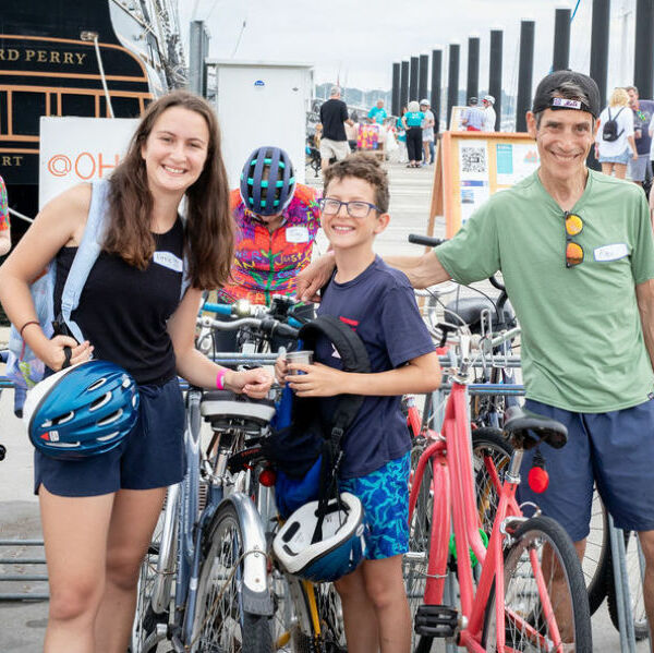 Family smiling with their bikes at the 10 spot ride. The stern of the Oliver Hazard Perry tall ship in the background