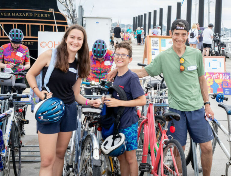 Family smiling with their bikes at the 10 spot ride. The stern of the Oliver Hazard Perry tall ship in the background