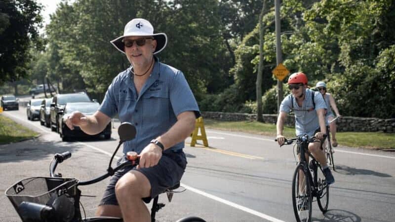 People riding bikes on a scenic road in Newport with lush greenery and cars in the background.