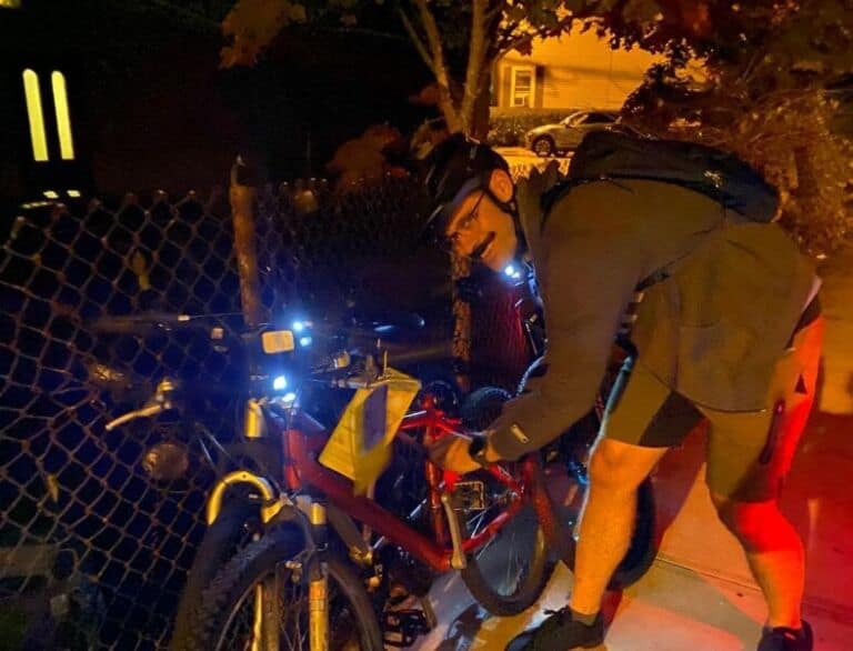 A cyclist locks bicycle at night with bike lights on, near a fence, wearing helmet and gear, illuminated by streetlight, in an urban setting for Bike Newport.
