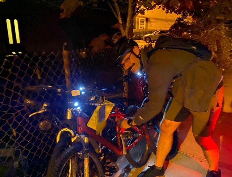 A cyclist locks bicycle at night with bike lights on, near a fence, wearing helmet and gear, illuminated by streetlight, in an urban setting for Bike Newport.