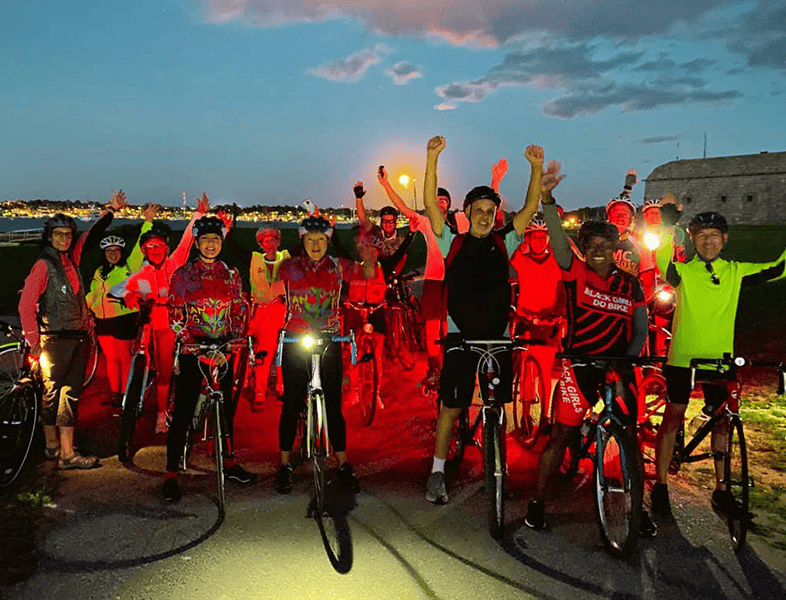 A diverse group of cyclists celebrating during a dusk bike ride along Newport waterfront, promoting community, fitness, and bike safety in Newport, Rhode Island.