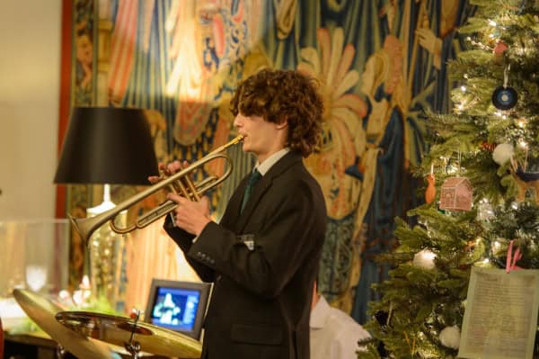 A young musician playing a trumpet during a festive holiday event with a Christmas tree and colorful curtains in the background.