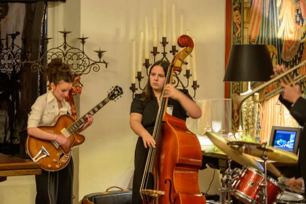 A jazz band performs live music indoors with a guitarist, bassist, and drummer at a cozy event space decorated with candles and artwork.