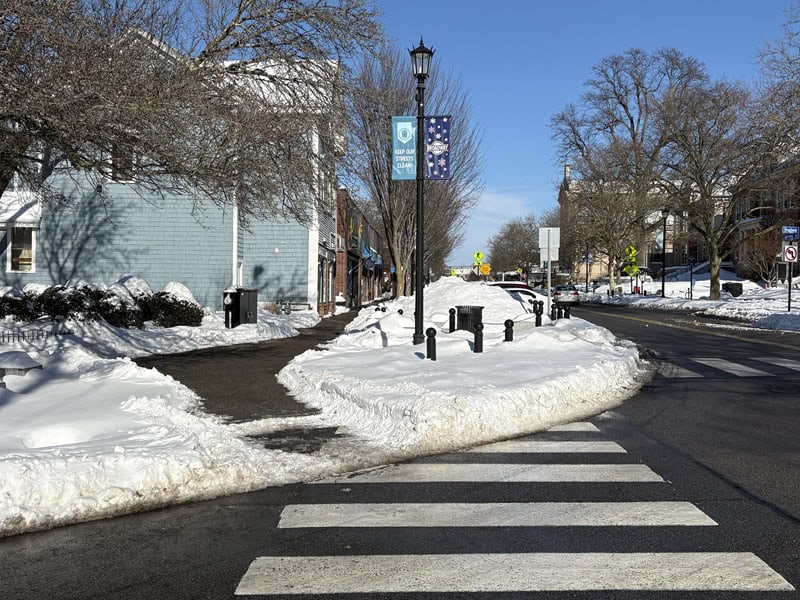A a snowy urban street in Newport, Rhode Island, featuring a sidewalk cleared for pedestrians and snow creating a sneckdown