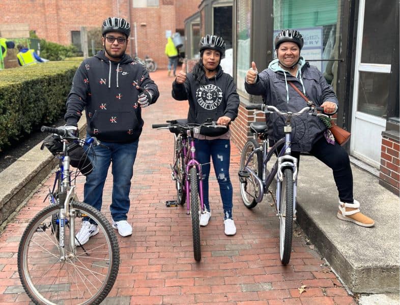 A diverse group of three cyclists, two women and one man, standing with their bikes on a city sidewalk, smiling and giving thumbs up, promoting inclusive cycling and community engagement.