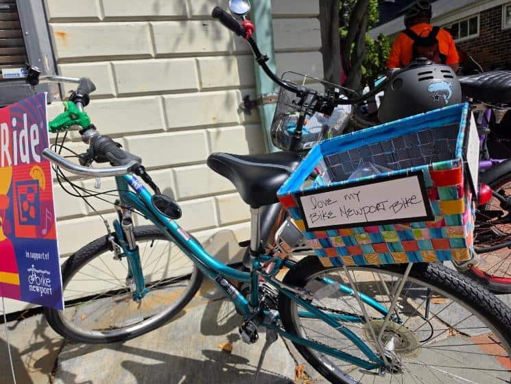 A teal bicycle with a colorful woven basket leans against a wall, a handwritten sign reading "I love my Bike Newport bike."
