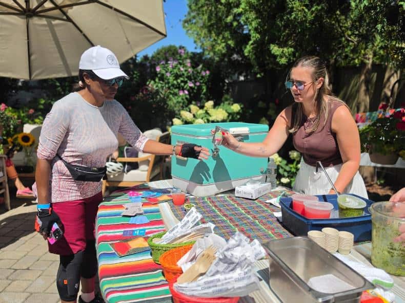 A server hands a drink across a serape-covered table to a rider in a white cap in a garden.