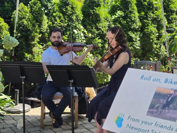 A violin duo performs seated in a garden beside a sign reading "Roll on, Bike Newport! From your friends at the Newport String Project."