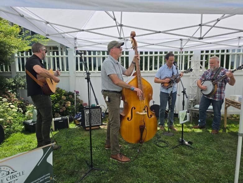 A four-piece string band with upright bass, mandolin, banjo and guitar performs under a white tent on a lawn.