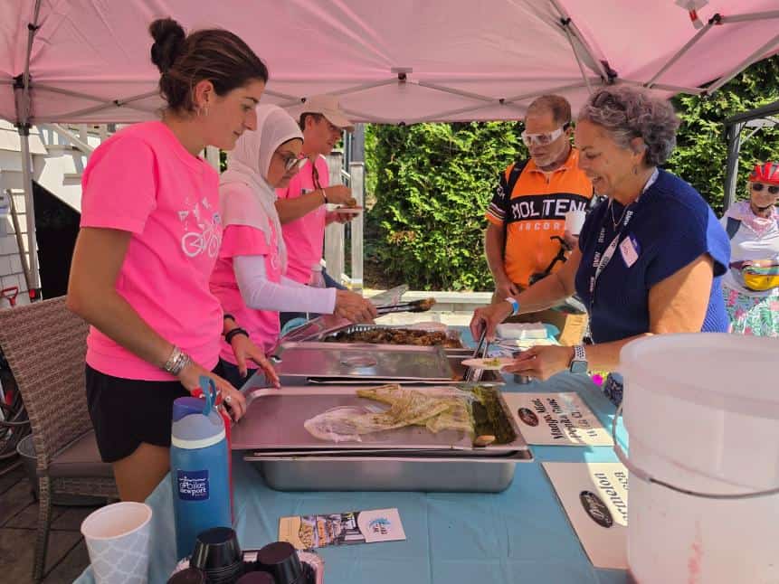 Volunteers in pink shirts plate food along a serving line under a pink tent as a rider in an orange jersey waits.