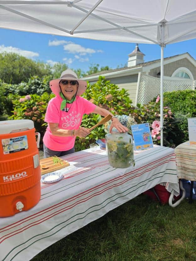 A volunteer in a pink shirt and sun hat smiles behind a drinks table with a cooler and a large jar of cucumber-mint water.
