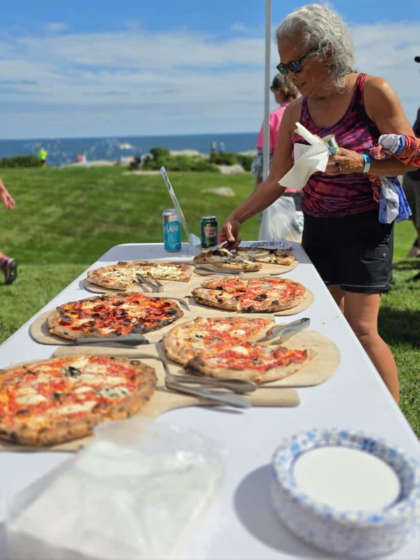 Six wood-fired pizzas on a long white table, a rider serving themselves a slice, the ocean behind.