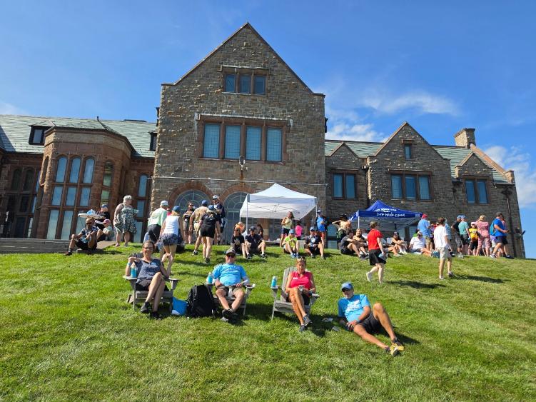 Riders relax in Adirondack chairs on a sloping lawn in front of a large stone mansion, event tents at the top.