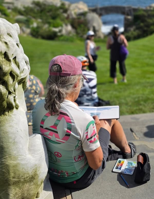 A rider seen from behind sketches with a watercolor set, resting against a stone sculpture as others gather on the lawn by the water.