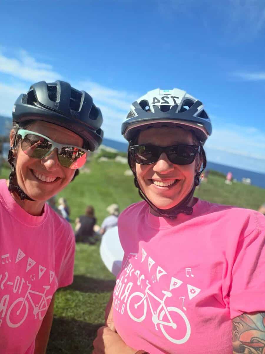 Two riders in pink shirts and bike helmets lean in for a close photo, the ocean behind them.
