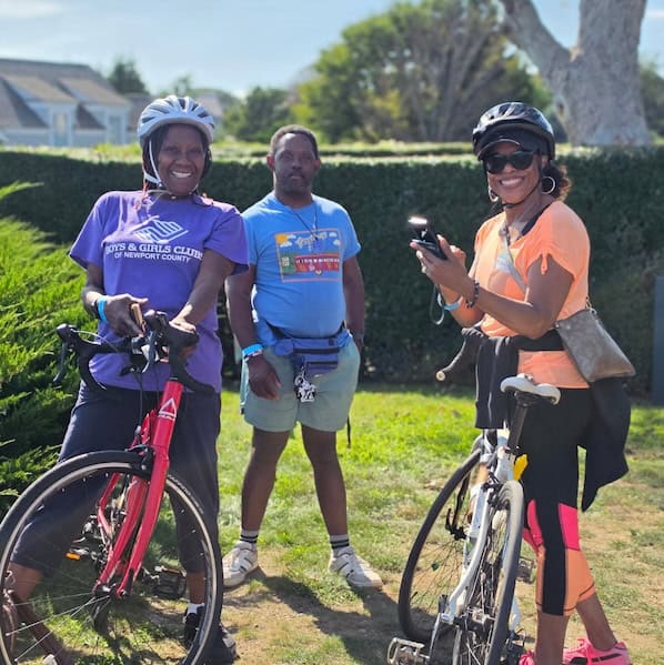 Three riders pause by a garden hedge with their bikes, one in a Boys & Girls Clubs of Newport County shirt, another holding a camera.