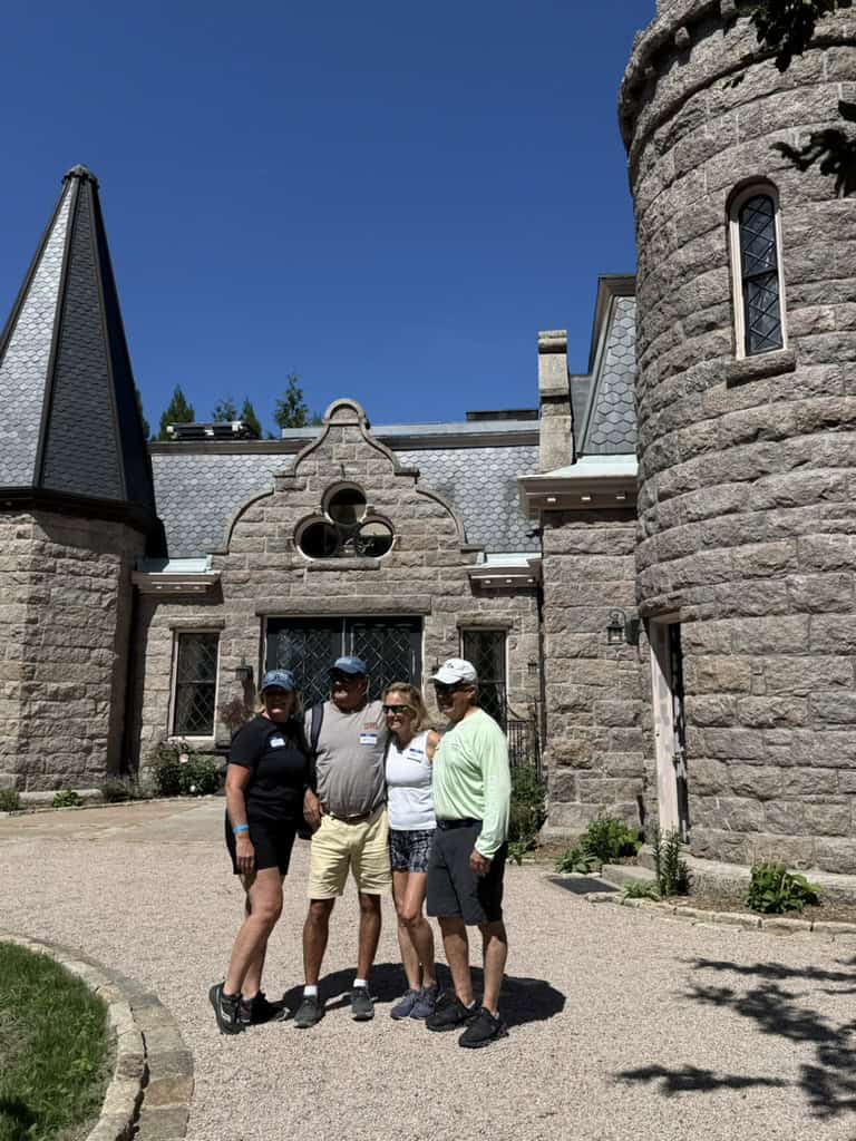 Four riders pose together in front of a stone carriage house under a bright blue sky.