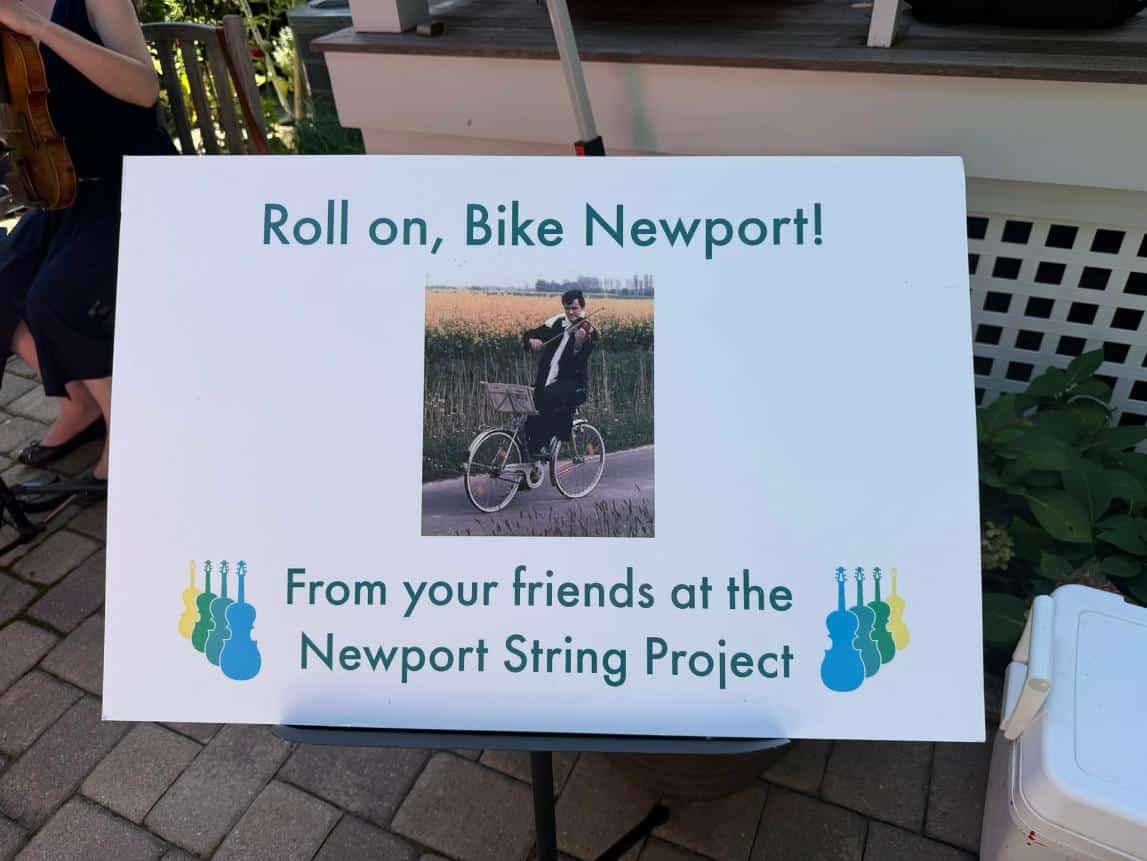 An easel sign reading "Roll on, Bike Newport! From your friends at the Newport String Project," with a photo of a cellist on a bicycle.