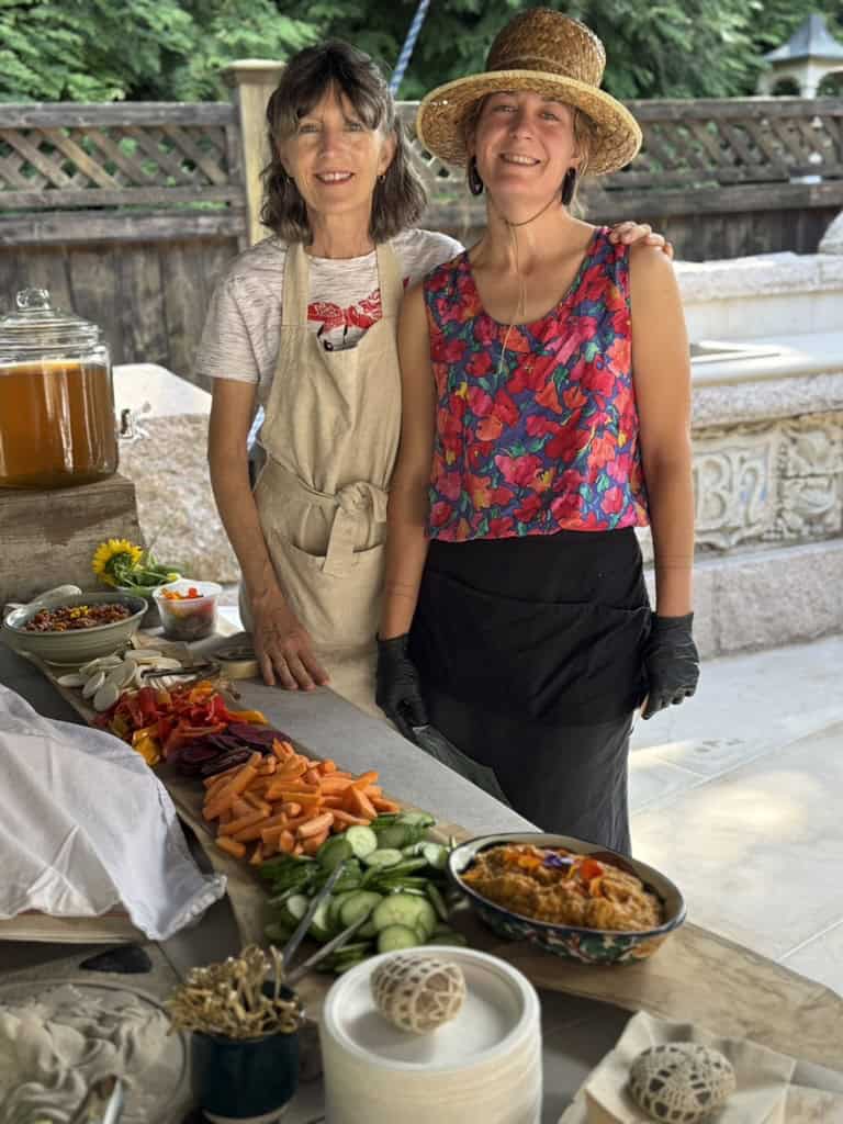 Two people stand behind a long grazing table of cut vegetables and dips, one in a straw hat and floral top, one in an apron.