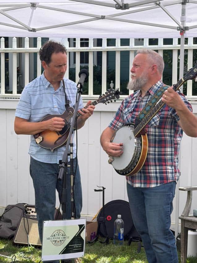 Two musicians from Millstone Circle play mandolin and banjo beside their band sign.