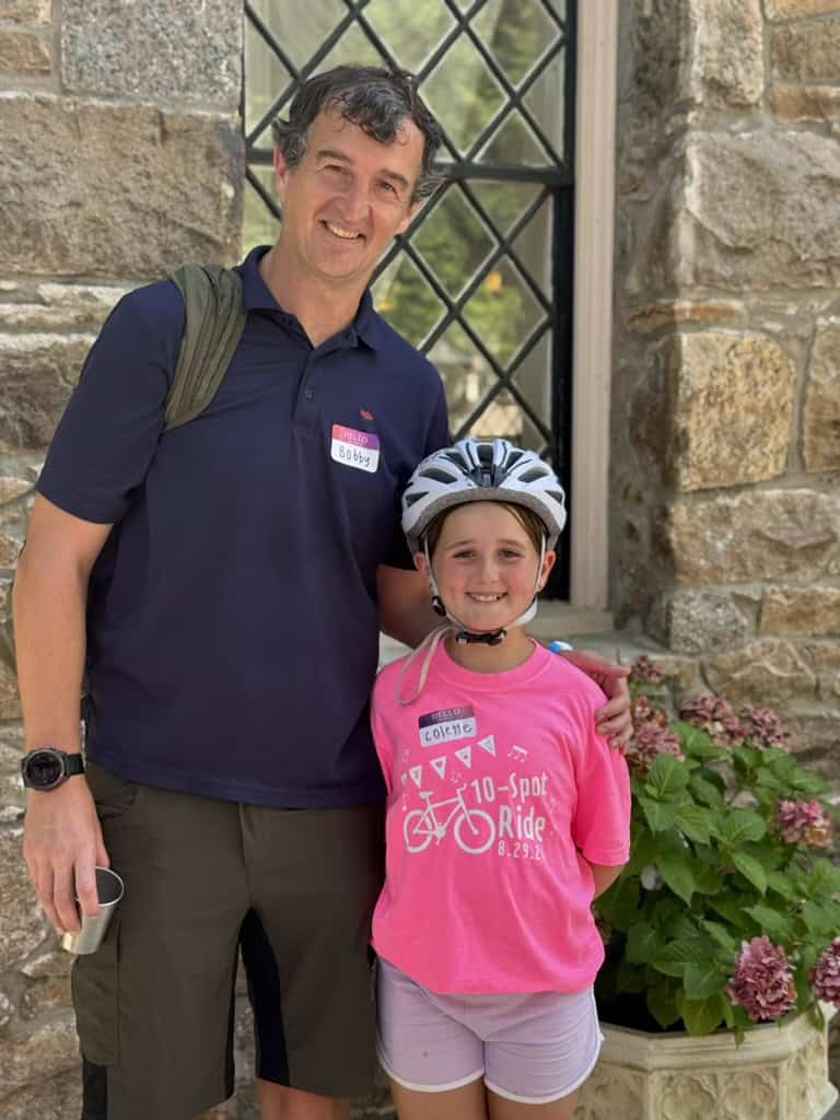 A man in a navy polo stands with a young rider in a pink 10-Spot Ride shirt and bike helmet outside a stone mansion window.