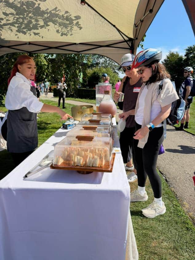 A caterer serves sandwiches to two helmeted riders at a white table beside a park path.