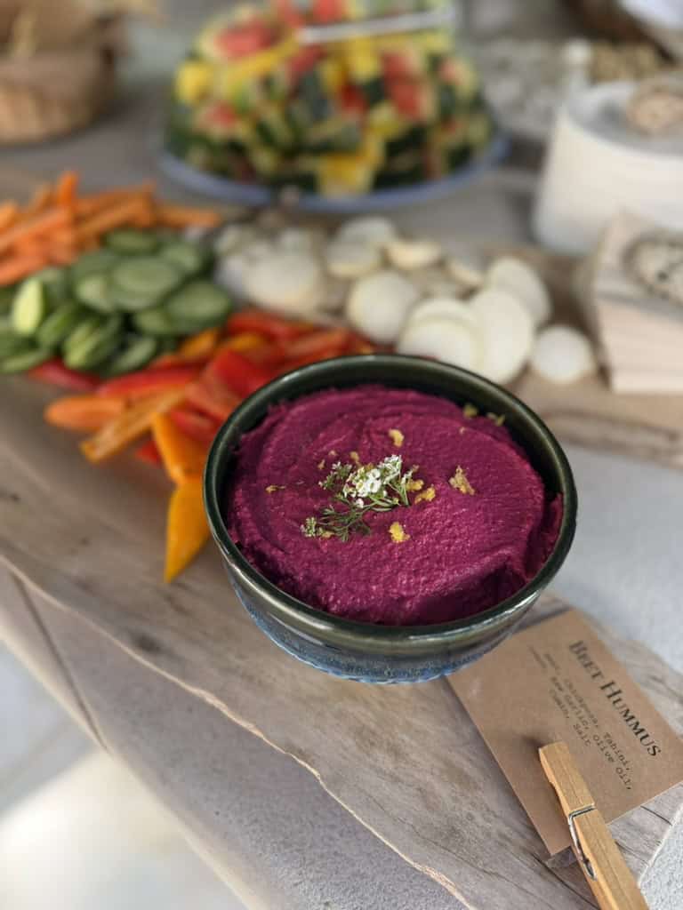 A bowl of magenta beet hummus garnished with herbs on a wood board with vegetable sticks and a "Beet Hummus" label.