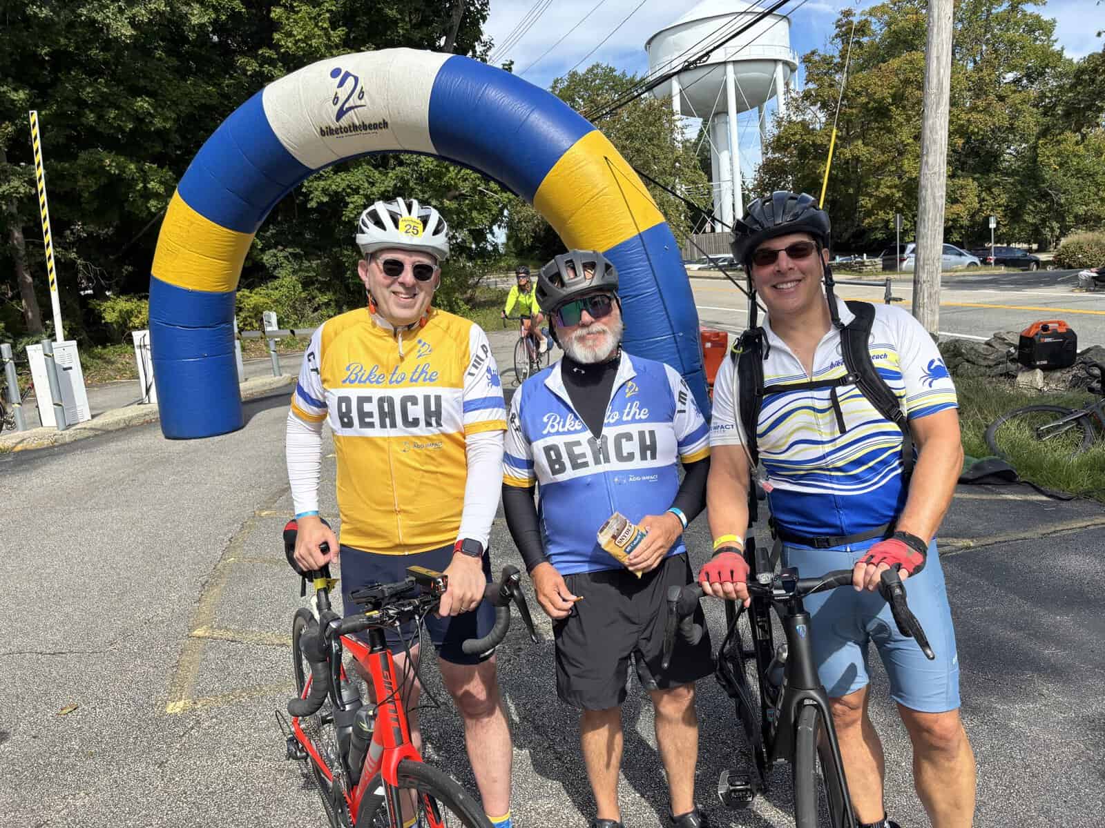 Three cyclists in Bike to the Beach jerseys standing under the inflatable arch in Woonsocket, Rhode Island, water tower in background
