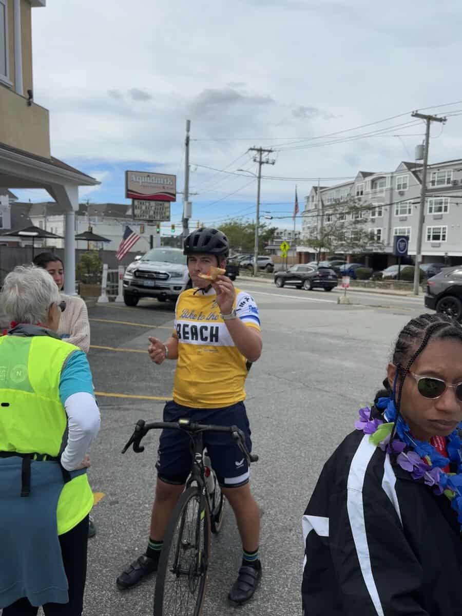 Cyclist in a yellow Bike to the Beach jersey eating a slice of pizza outside Aquidneck Pizzeria in Middletown