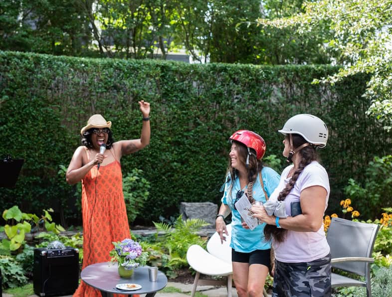 A performer in an orange dress sings into a microphone for two helmeted riders in a private garden at the 10-Spot Ride.