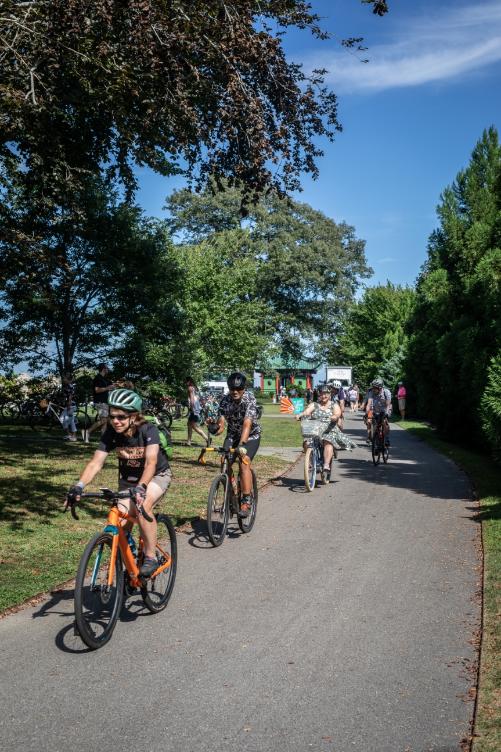 A line of riders travels a tree-shaded park path, the lead rider on an orange bike.