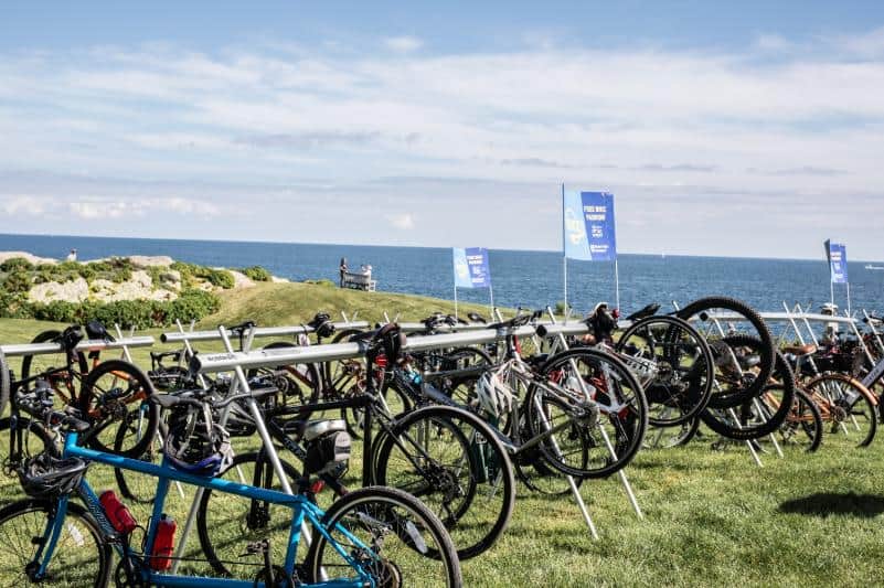 Rows of bicycles in valet parking racks on a lawn above the ocean, blue "free bike valet" flags flying.
