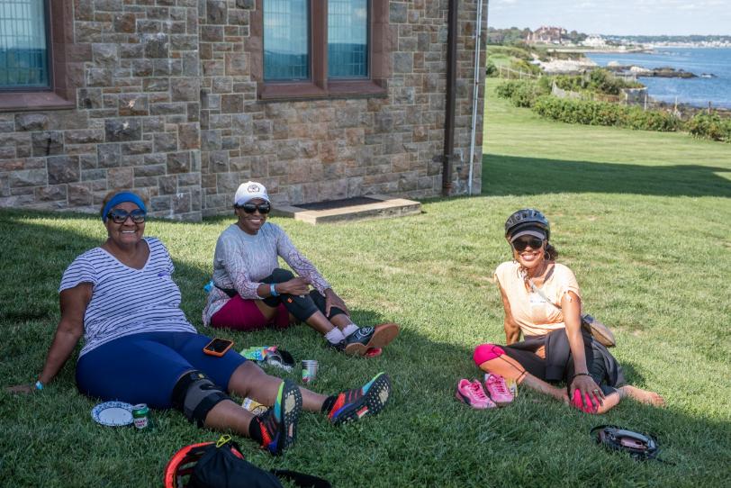 Three women sit together on a lawn beside a stone mansion, smiling, the ocean behind them.