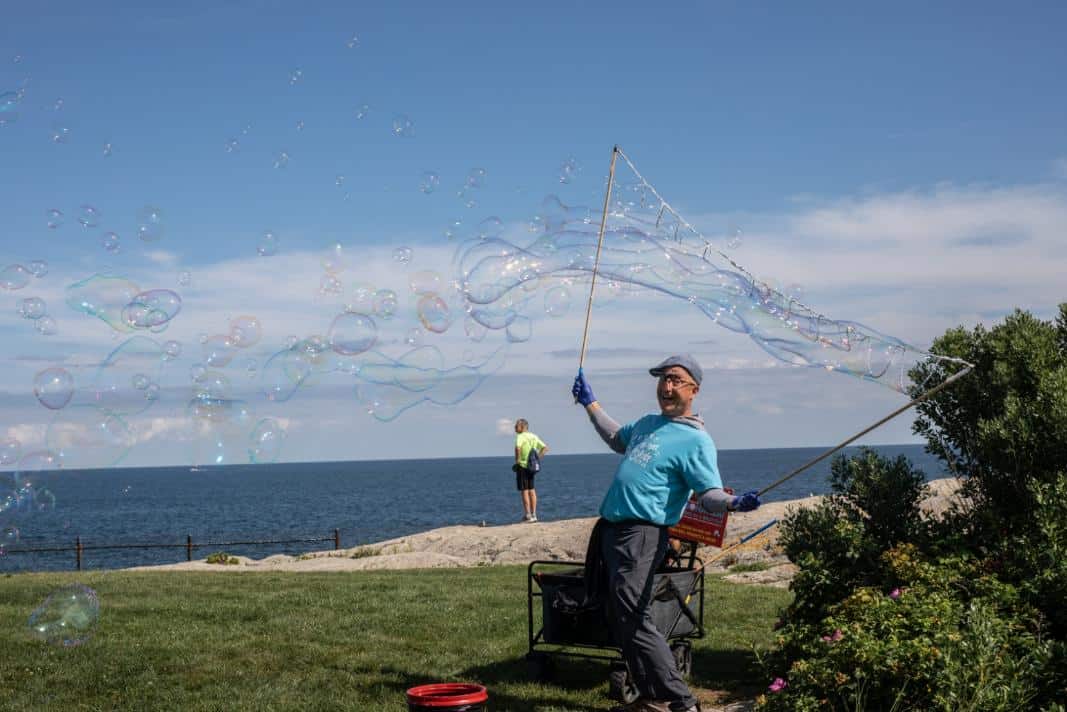 A performer draws a long stream of giant soap bubbles with two wands on a lawn above the ocean.