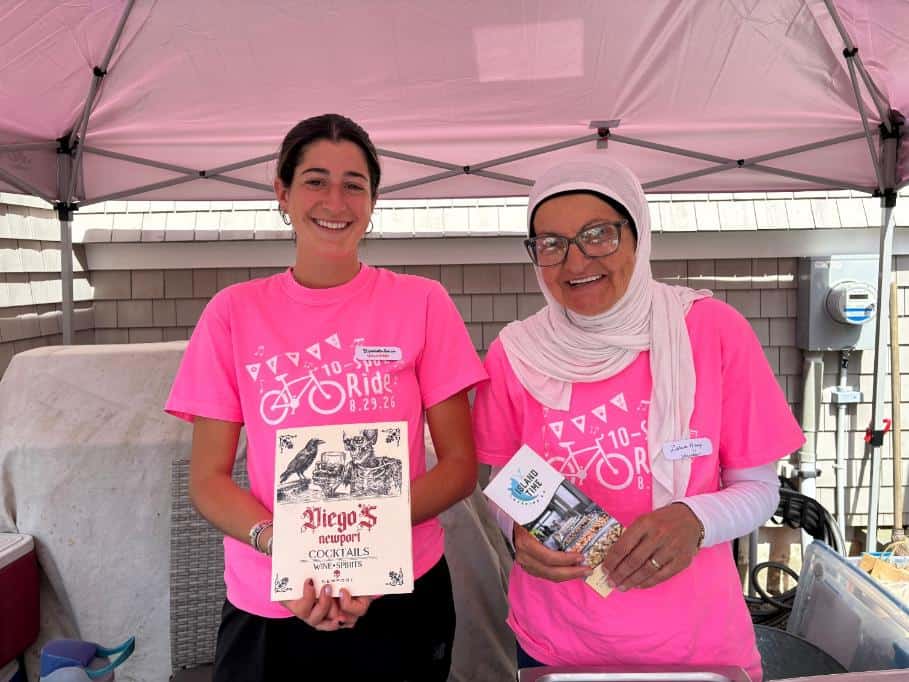 Two volunteers in pink shirts under a tent, one holding a printed Diego's Newport cocktails sign.