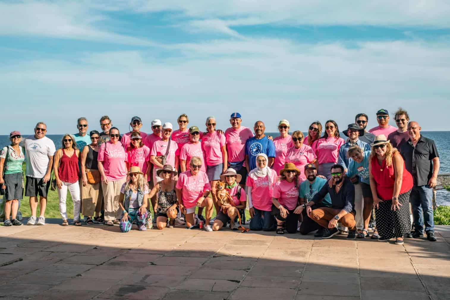 About forty staff and volunteers in pink shirts pose for a group photo on a stone terrace above the ocean.